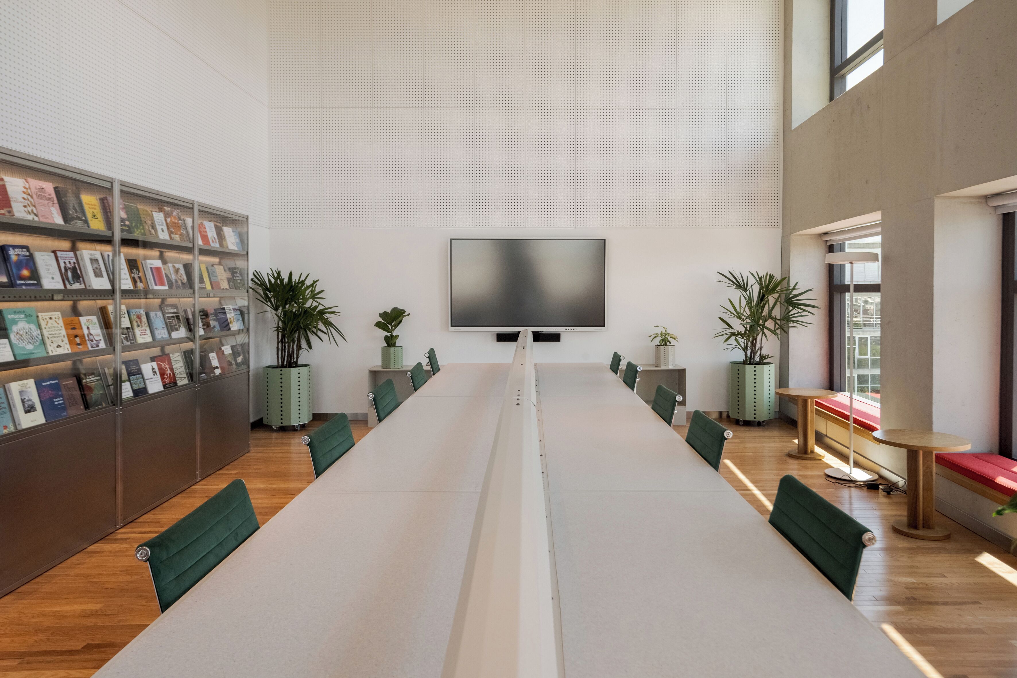 A spacious library with a long table, green chairs, a large bookshelf, and a flat-screen TV on the wall.