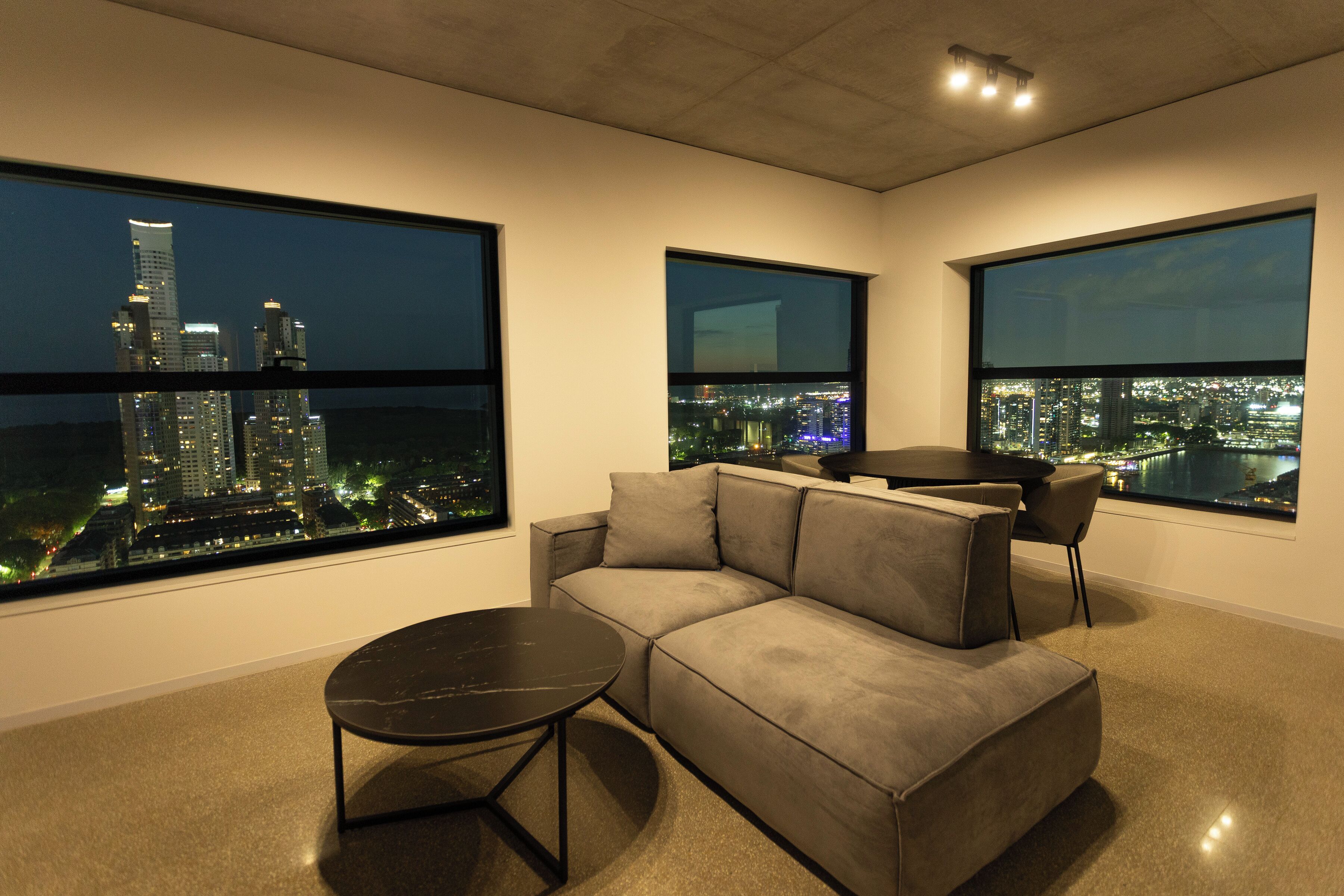 Uma sala de estar moderna com um sofá cinzento, uma mesinha redonda e janelas amplas que oferecem vista noturna da cidade.