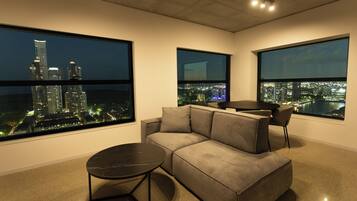 Uma sala de estar moderna com um sofá cinzento, uma mesinha redonda e janelas amplas que oferecem vista noturna da cidade.