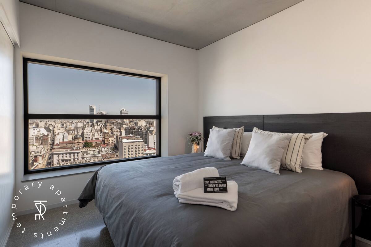 A modern hotel room with a large window, a neatly made bed, and a city view.