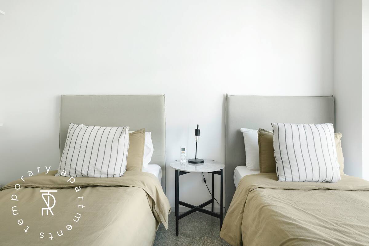 A hotel room with two beds, a small table with a lamp, and a plain wall.