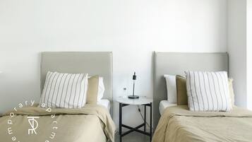 A hotel room with two beds, a small table with a lamp, and a plain wall.
