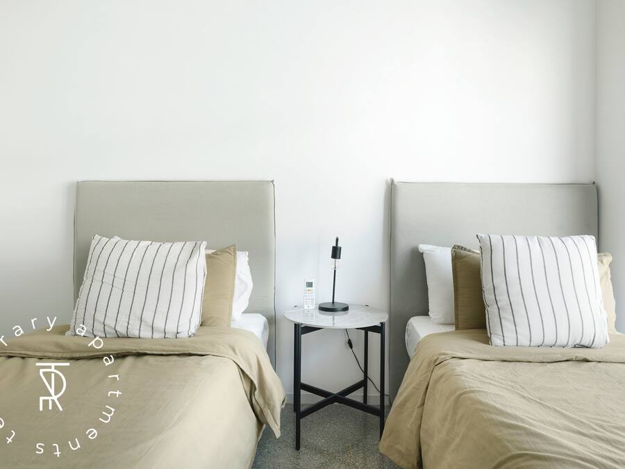A hotel room with two beds, a small table with a lamp, and a plain wall.