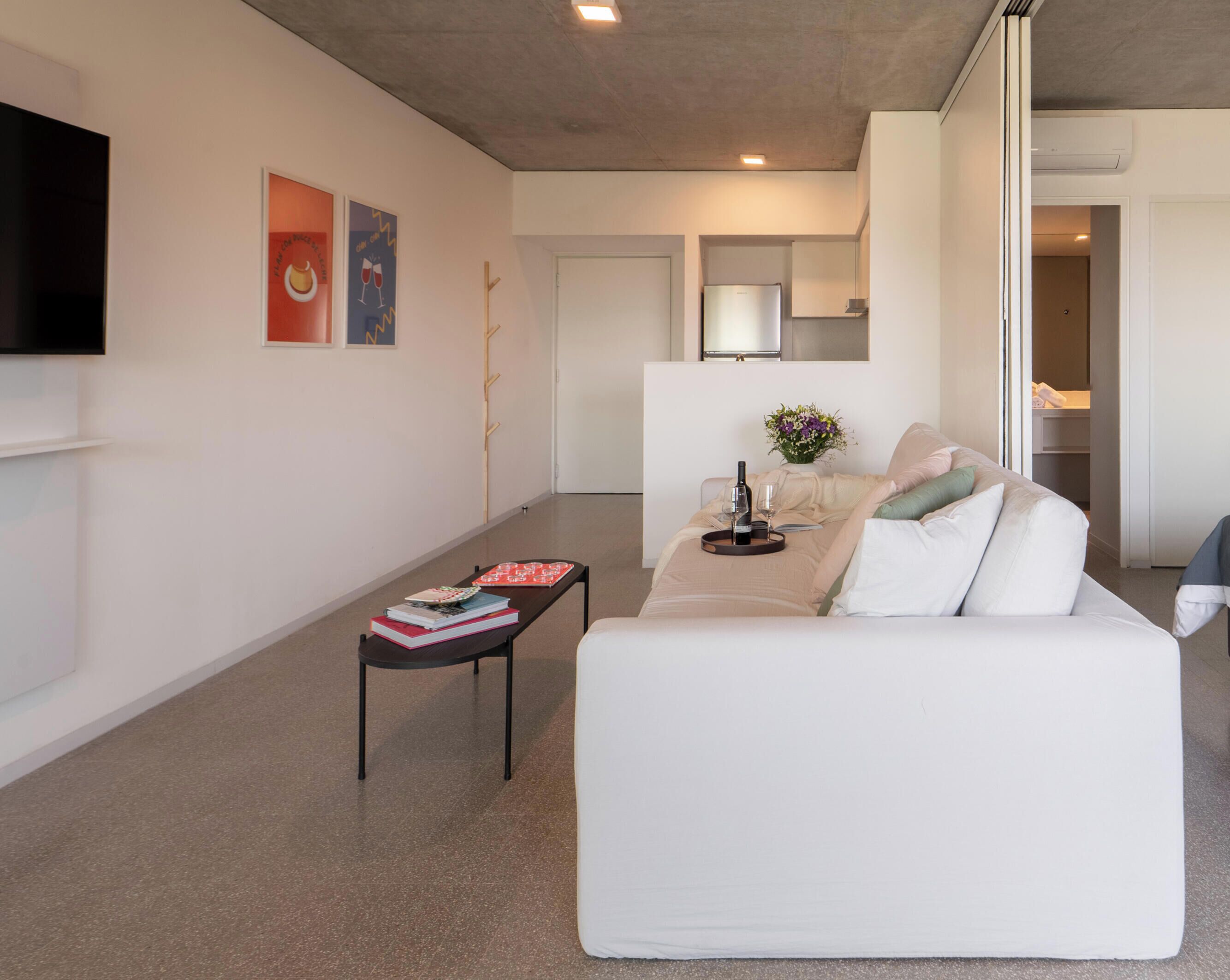 Un salon moderne avec un canapé blanc, une table basse sur laquelle sont posés des livres, un téléviseur à écran plat fixé au mur et, au fond, un coin cuisine.