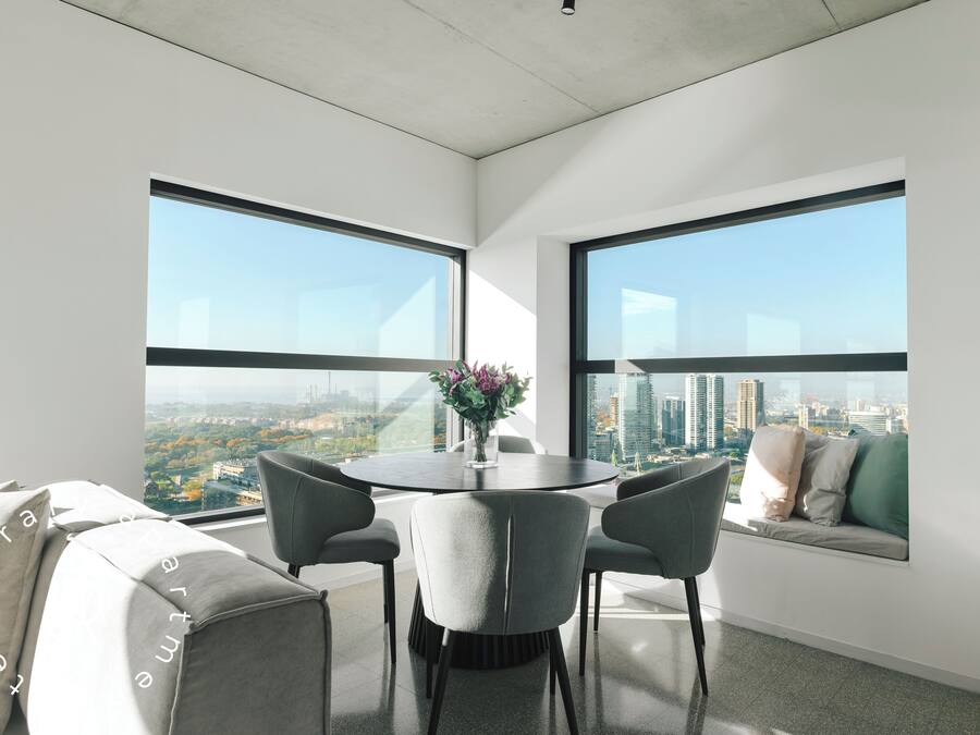 Een moderne kamer met een eettafel, grijze stoelen, een bank met kussens en grote ramen met uitzicht op de stad.