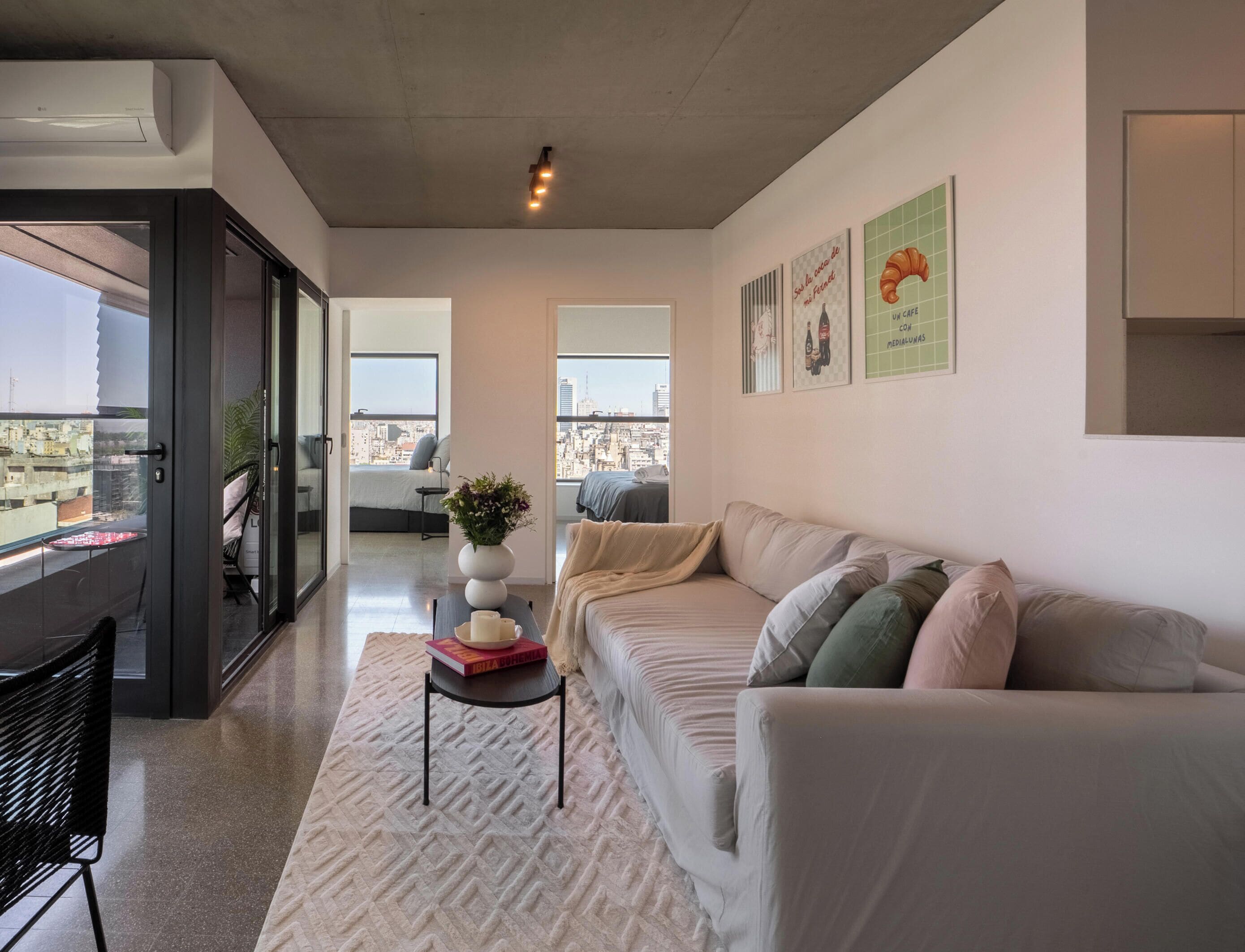 Una sala de estar moderna con un sofá, una mesita con libros, un jarrón decorativo y vistas a la ciudad a través de amplios ventanales.