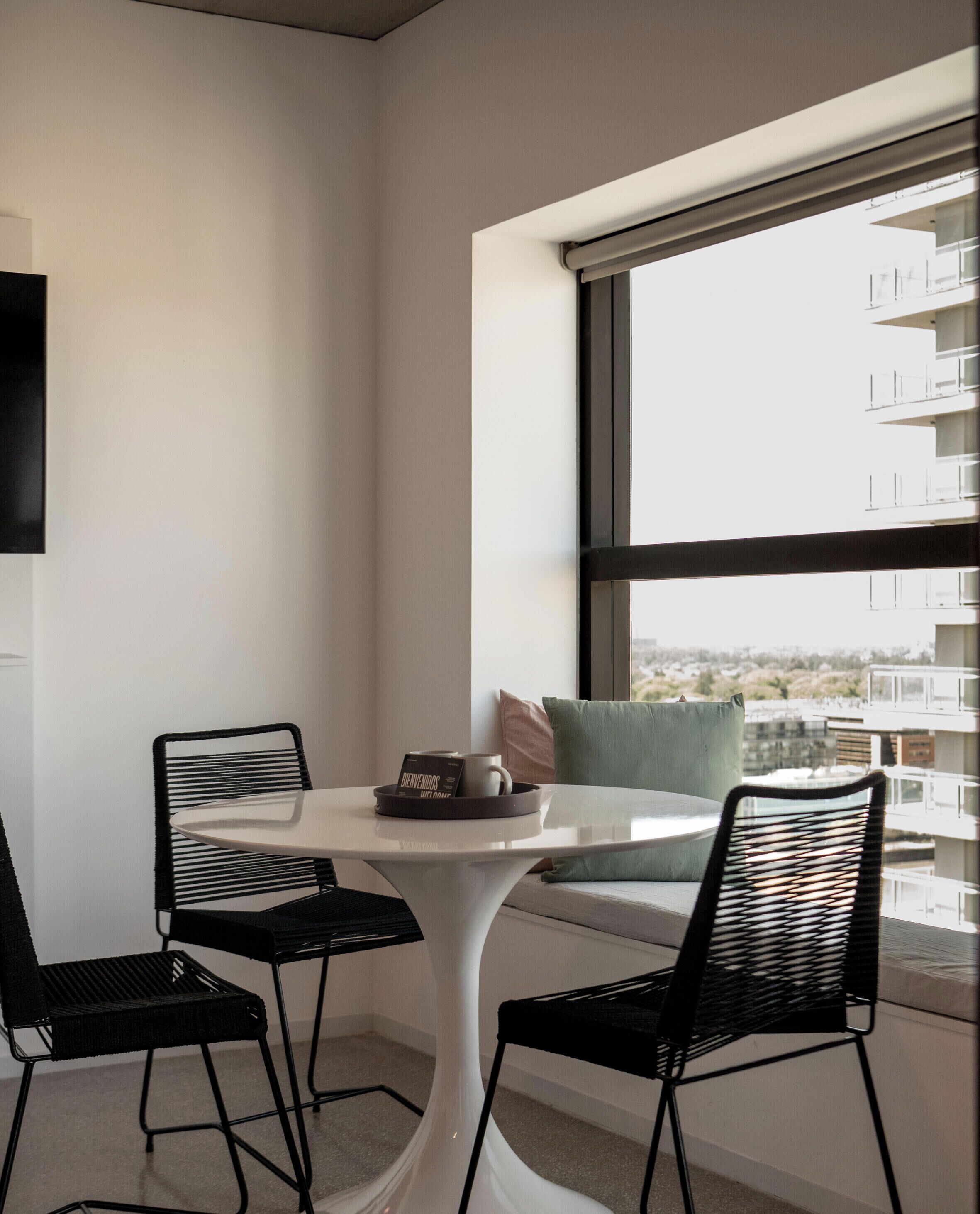 A modern room with a round table, two black chairs, a window with a view of buildings, and a white wall.