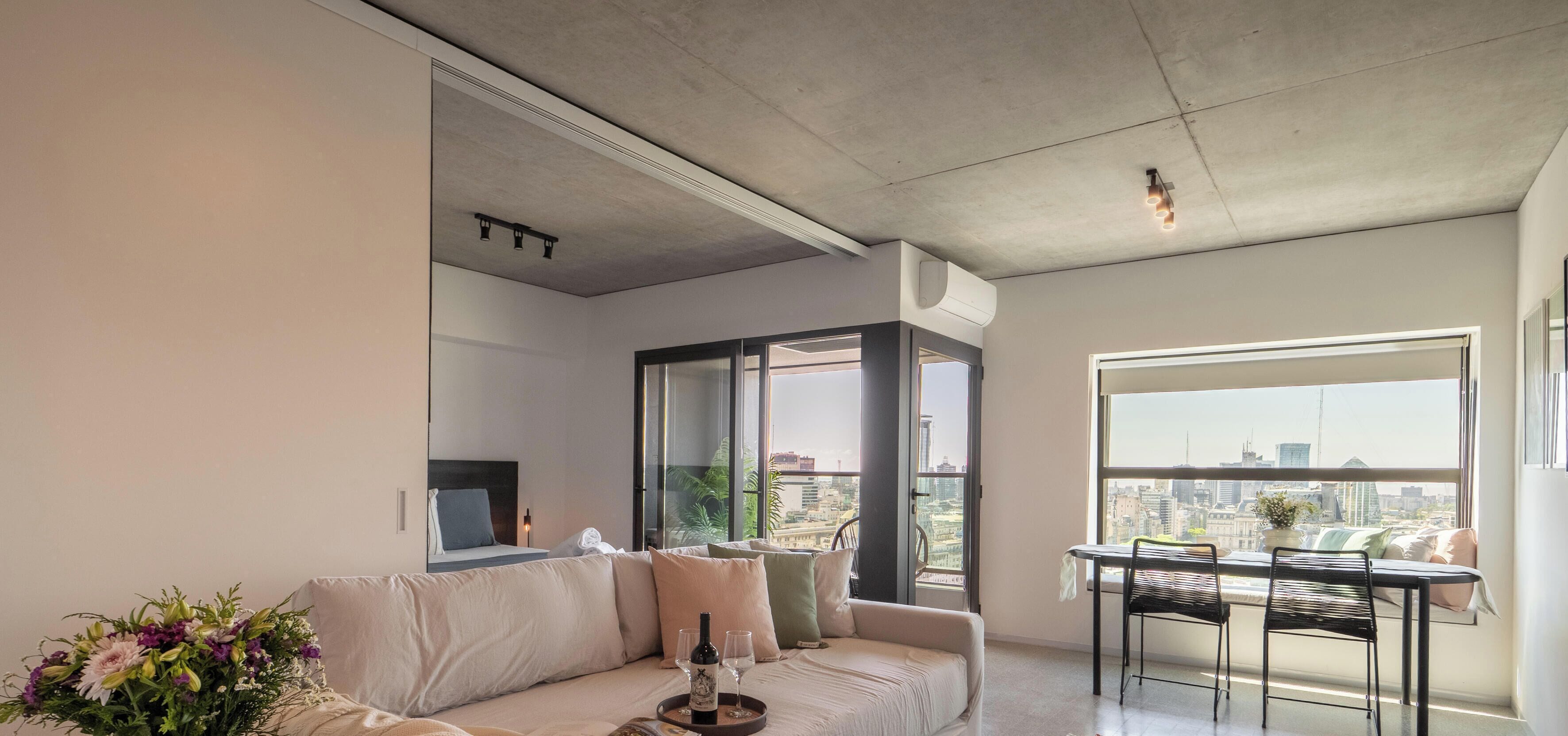 A modern living room with a sofa, a dining table, and a large window offering a city view.