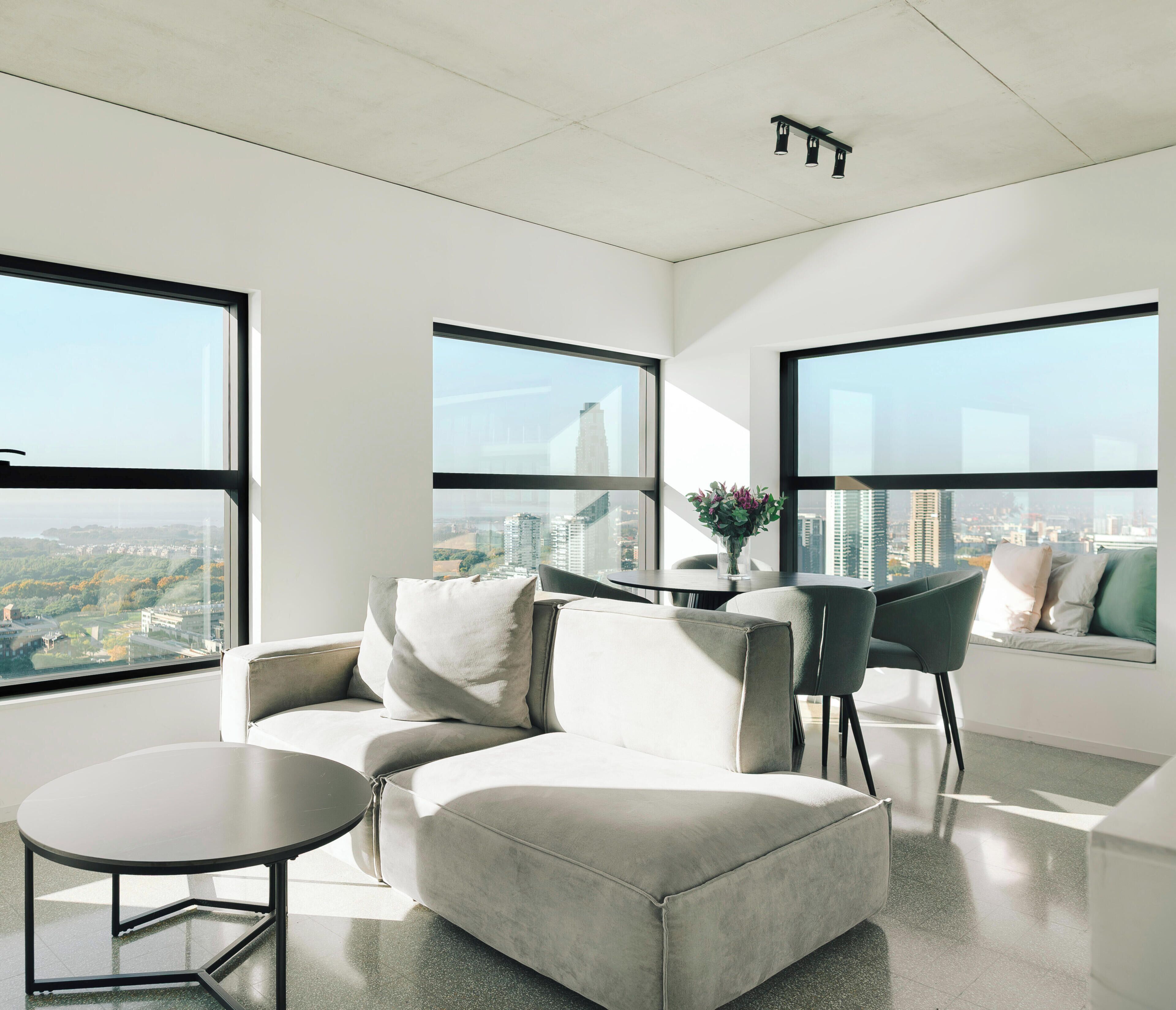 Un salon moderne avec un canapé, une petite table et un fauteuil. De grandes fenêtres offrent une vue sur la ville.