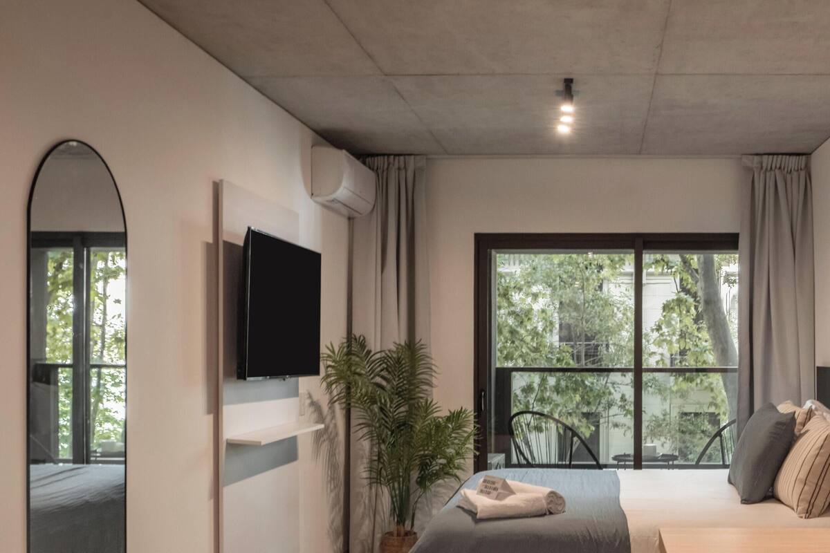 A modern bedroom with a bed, a TV, a potted plant, a window with a view of trees, and a decorative vase.