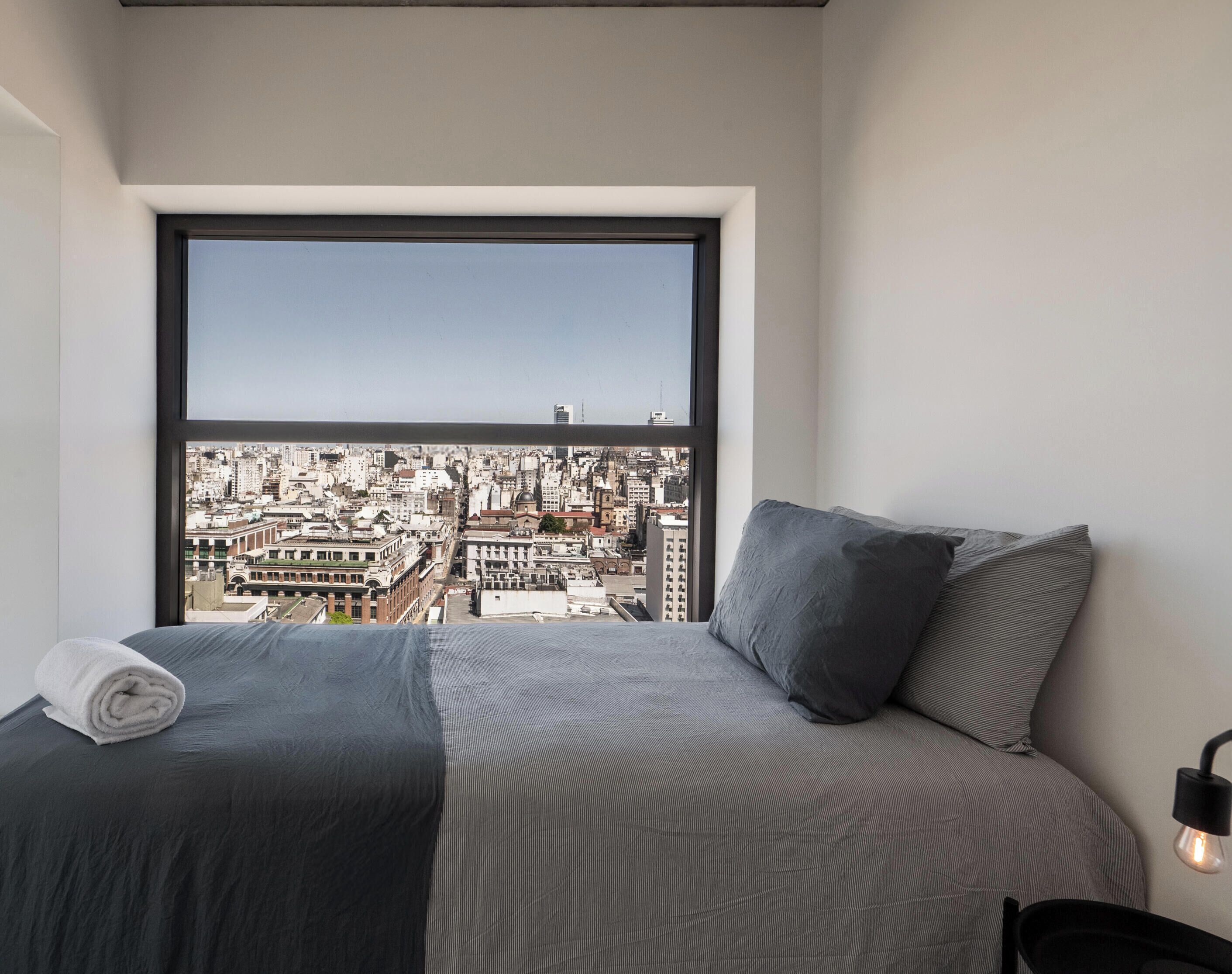 A bedroom with a large window, a bed with grey bedding, and a city view.