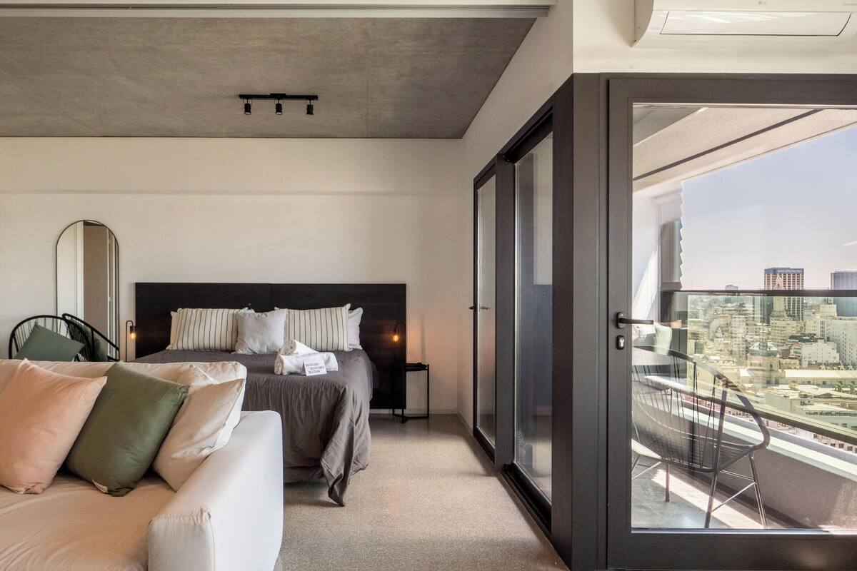 A modern hotel room with a large bed, a sofa, a nightstand, and a balcony with a city view.