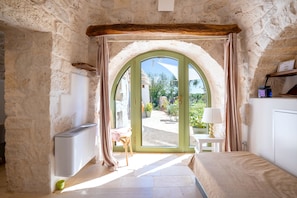 A room with a stone wall, a bed, a small table with a lamp, and a view through an arched doorway to an outdoor area with plants.