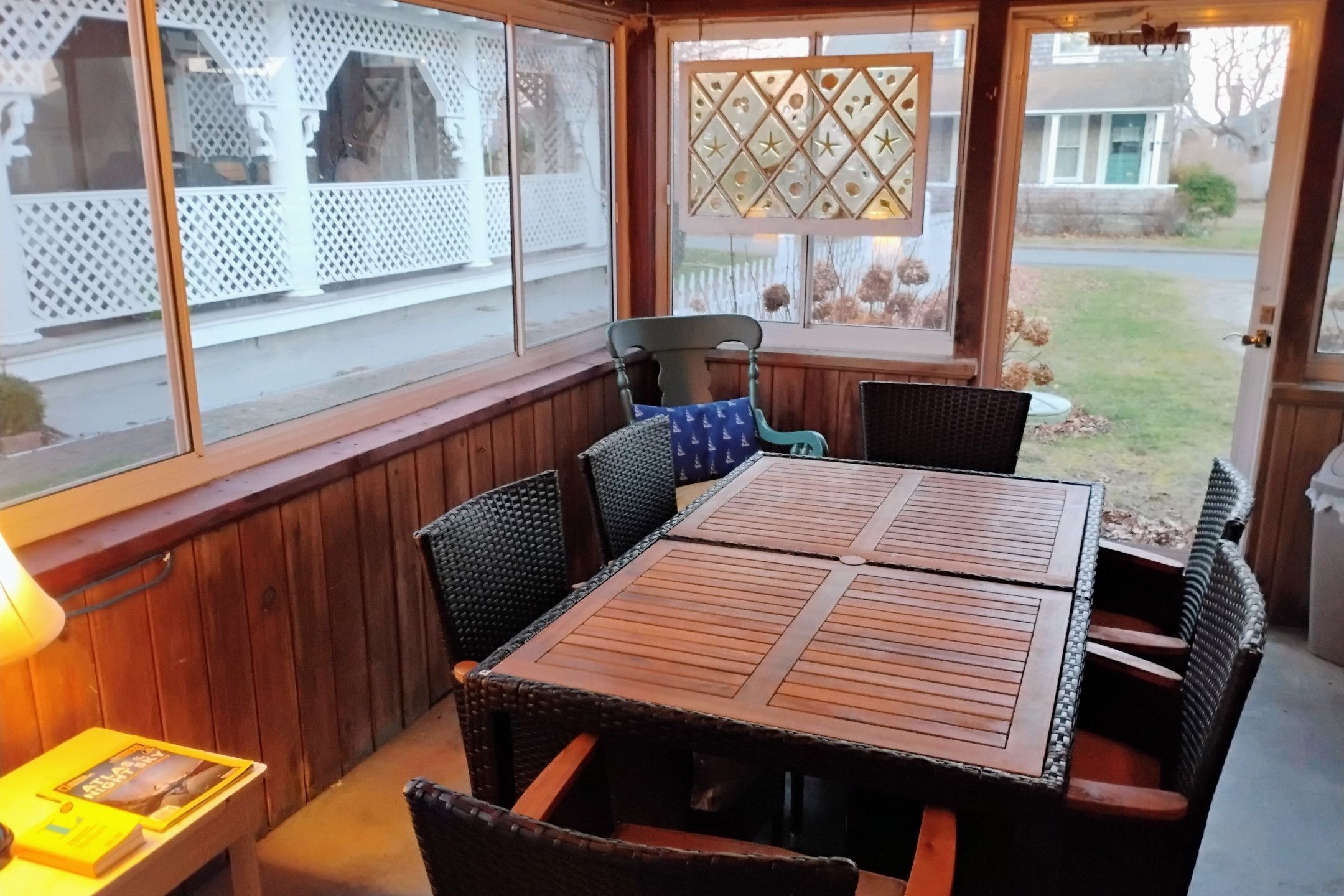 A covered patio with a wooden table and wicker chairs, a window with a lattice design, and a view of a residential area.