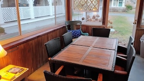Een overdekt terras met een houten tafel en rieten stoelen, een raam met een gesneden raamrooster en uitzicht op een woonwijk.