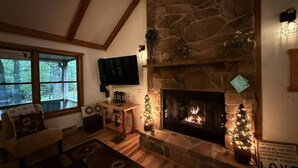 A cozy living room with a stone fireplace, a television mounted above it, and a Christmas tree.