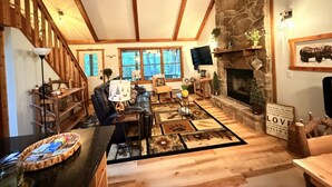 A cozy living room with a stone fireplace, wooden beams, a fireplace mantel, and a large window.