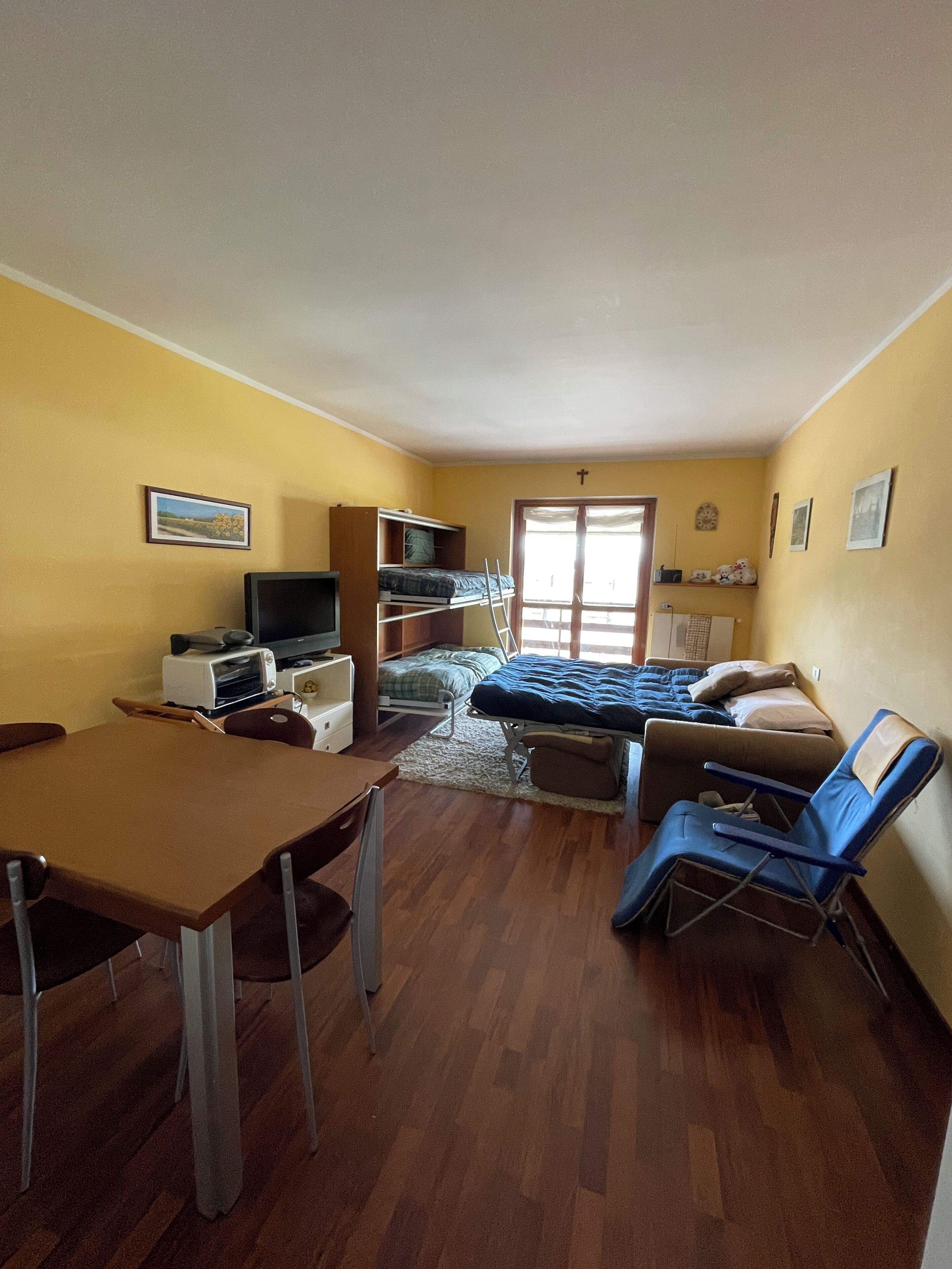 Een ruimtelijk ingerichte leefruimte met een bed, een televisie, een eettafel en een kitchenette.