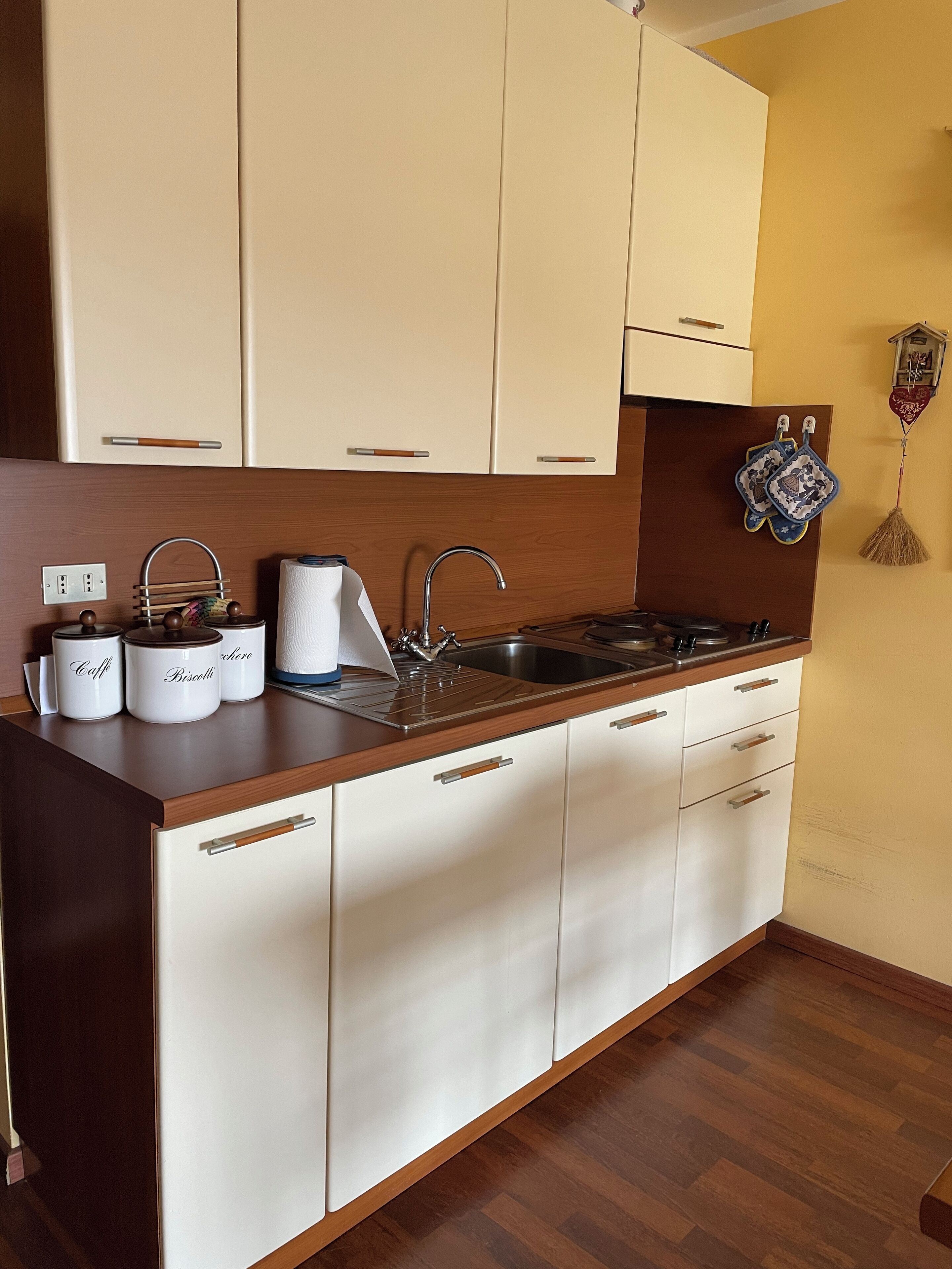 Een keuken met witte kastjes, een houten aanrecht, een roestvrijstalen spoelbak en een kookplaat. Drie witte bakken zijn gelabeld met 'Caffè', 'Biscegli' en 'Chicco'.