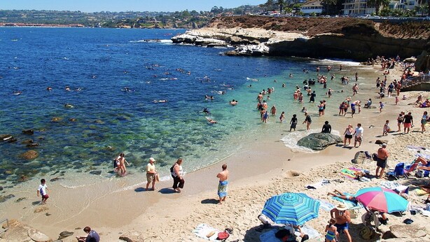 Une plage bondée, avec une eau claire, des gens qui se baignent et des parasols.