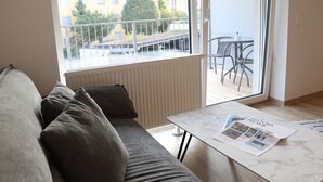 A modern living room with a sofa, a coffee table with magazines, a view of a balcony with chairs, and a window overlooking a building. - Comfort Apartments Villach Self CheckIn (Villach)