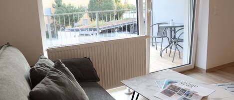 A modern living room with a sofa, a coffee table with magazines, a view of a balcony with chairs, and a window overlooking a building.