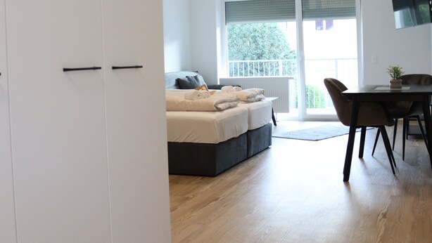A modern bedroom with a bed, a dining area with a table and chairs, and a large wardrobe. - Comfort Apartments Villach Self CheckIn (Villach)