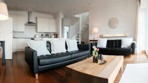 Een moderne woonkamer met een leren zwarte bank, een houten salontafel en een zwarte bank met witte kussens. In de achtergrond is een keuken met witte kasten en een trap te zien.