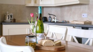Een eettafel met een fles BLANC PESCAO wijn, een glas, een potje olijven, een brood en een roze tulp.