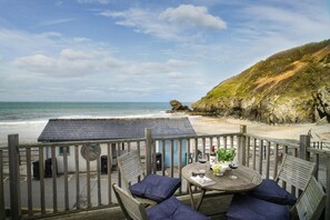 A wooden deck with a table and chairs overlooking a beach with a rocky cliff and ocean view. - Beach Hut Apartment, Llangrannog (Llangrannog)