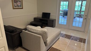 Een woonkamer met een grijze bank, een zwart voetenbankje, een tv op een houten dressoir, en een deur die naar buiten leidt.