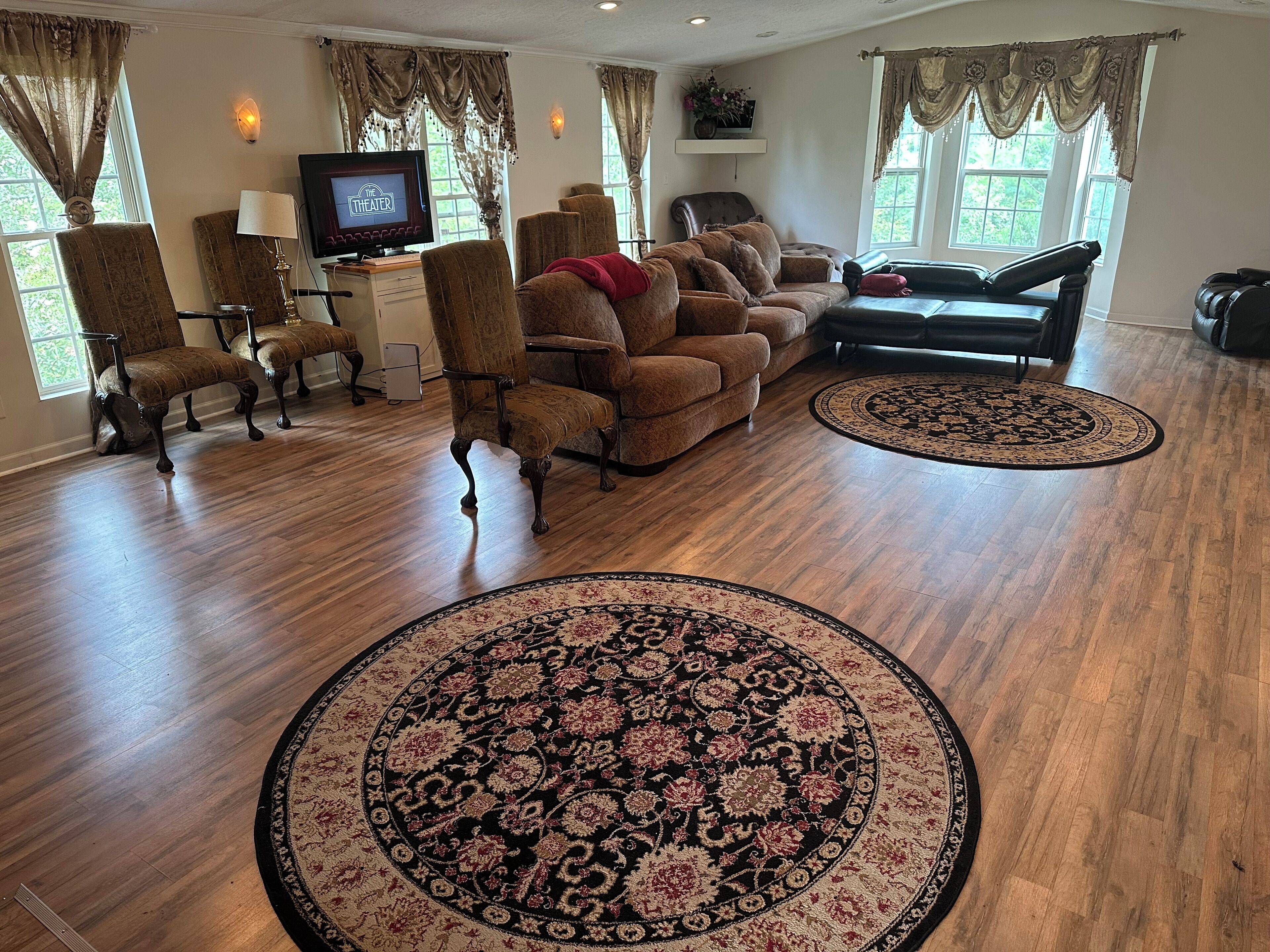 A spacious living room with a wooden floor, a large sofa set, two armchairs, a television, and a decorative rug.