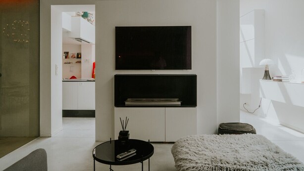A modern living room with a flat-screen TV mounted on the wall, a small round table with a remote control, and a fluffy white ottoman.