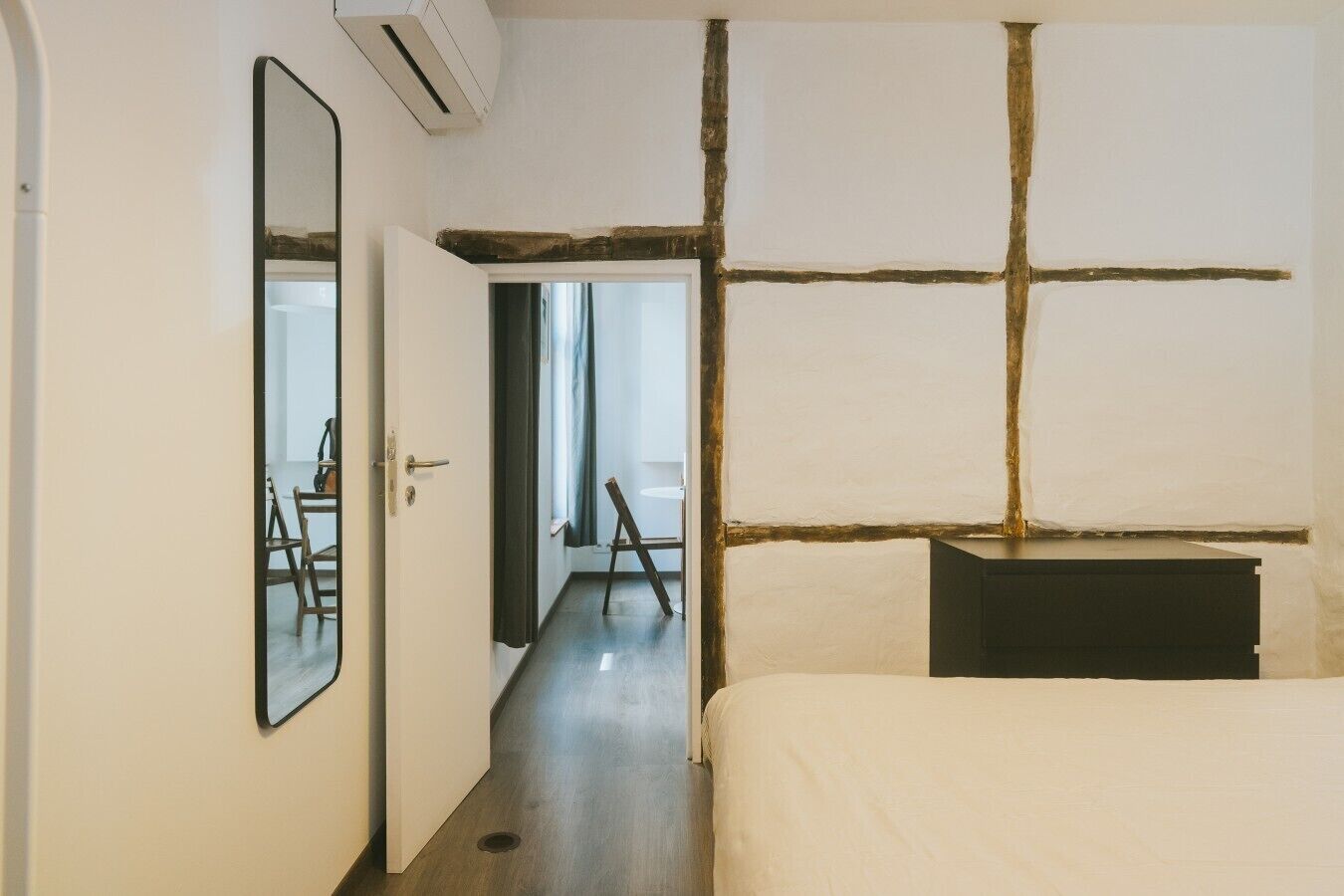 Een hotelkamer met een bed, een bureau, een stoel en een spiegel. Er is een deur die naar een andere kamer leidt.