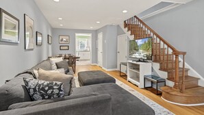 A modern living room with a grey sofa, a wooden staircase, and a TV on a white stand. - The Bella Vista Townhouse (Philadelphia)