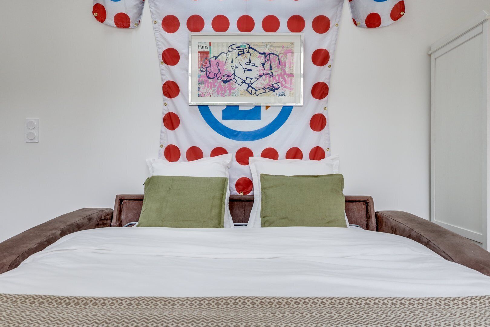Uma cama com um cobreleito estampado, duas almofadas verdes e uma parede com um desenho circular e um quadro emoldurado.