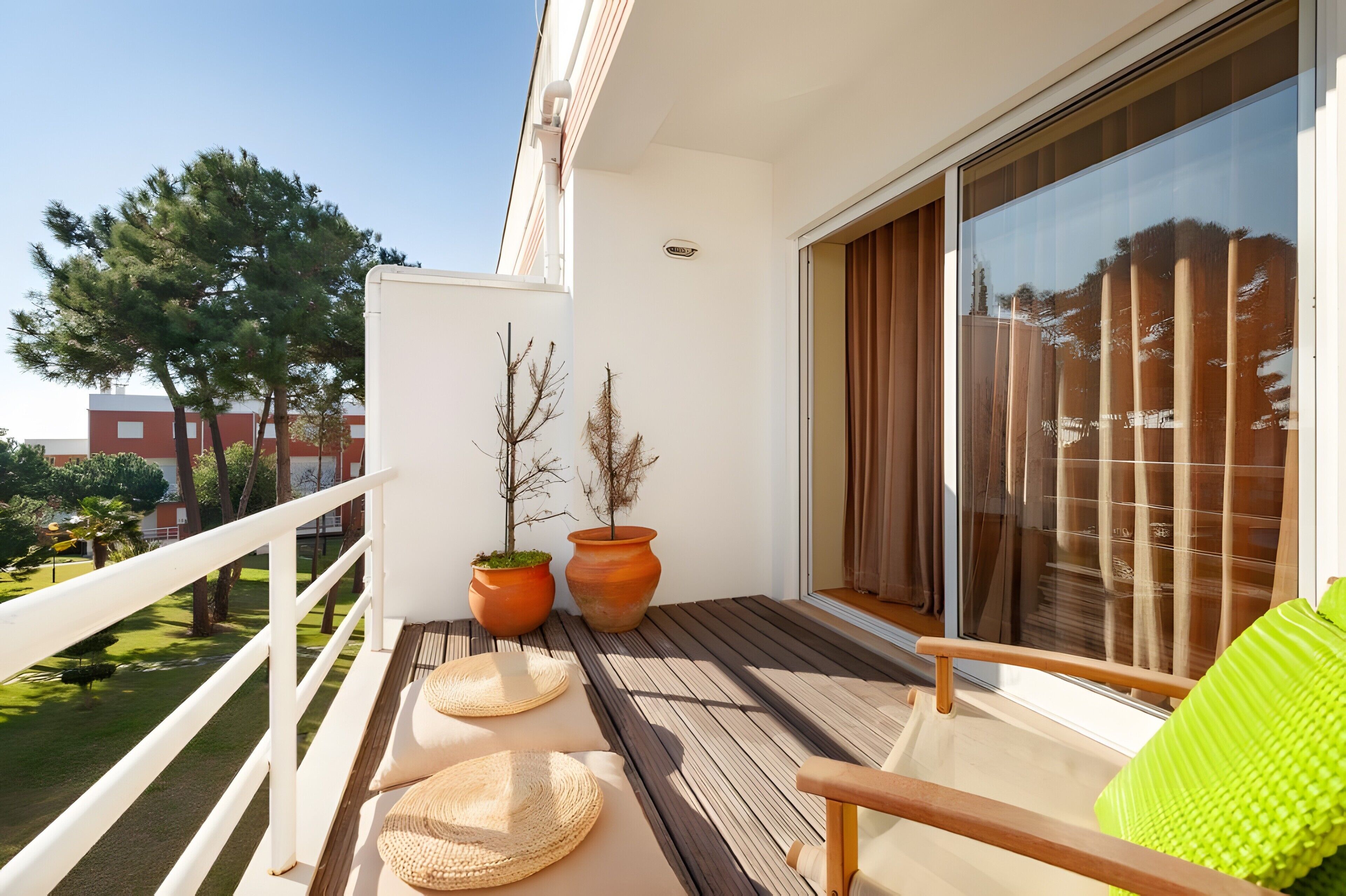 A balcony with a wooden floor, two potted plants, a chair with a green cushion, and a large window with curtains.