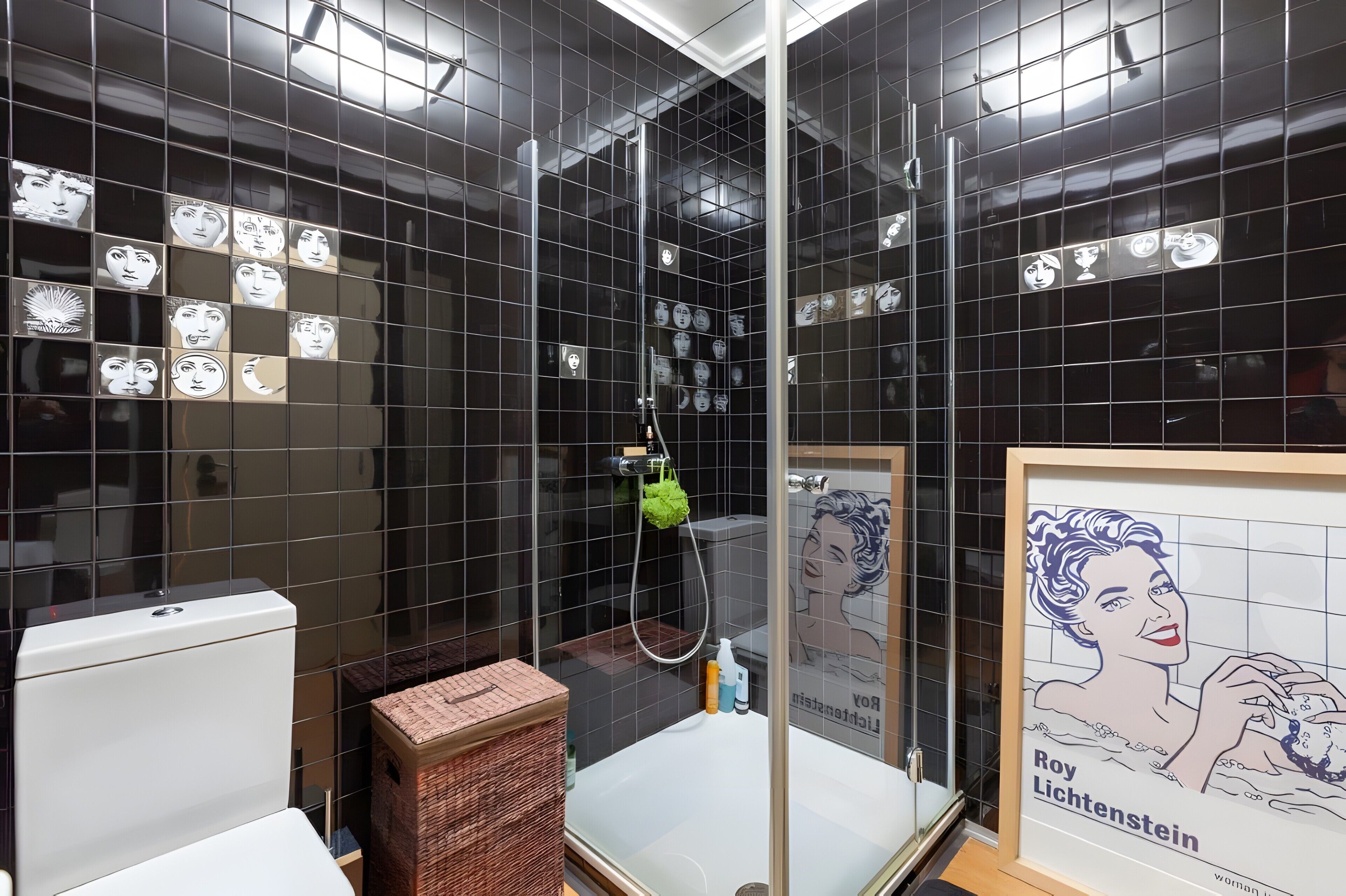 A bathroom with black tiled walls, a shower, a toilet, and a framed poster of Roy Lichtenstein.