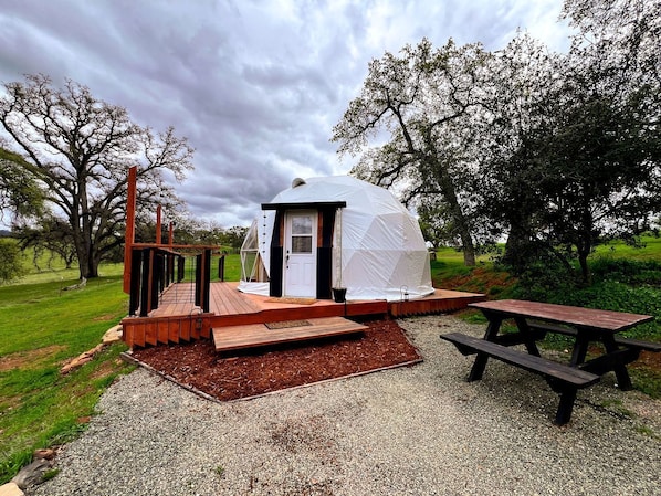 A small, dome-shaped tent with a door and windows, a wooden deck, and a picnic table.