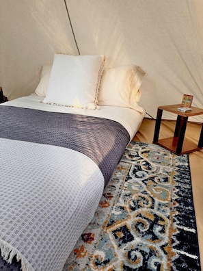 Un lit recouvert d’une couverture blanche et grise, un tapis à motifs et une petite table de chevet en bois sur laquelle repose un livre.