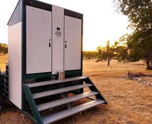 A large, white, metal structure with 'ECOZOIC BIOFILTER TOILET' text, a metal staircase, and a grassy field with trees in the background.
