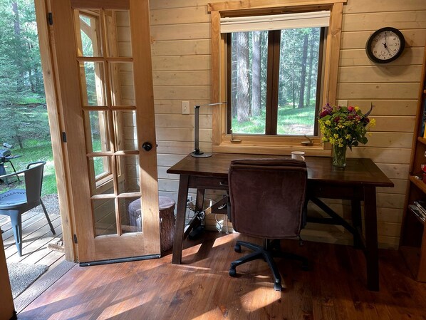 A wooden cabin interior with a desk, chair, and a window overlooking a forest.