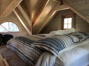 Un dormitorio acogedor en el ático, con techo inclinado, una cama con ropa de cama a rayas y un ventanal con vistas a los árboles.
