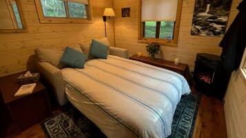 A cozy log cabin bedroom with a bed, a nightstand, a lamp, a window, and a fireplace.