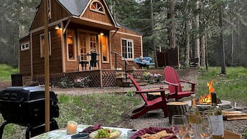 Eine Holz-Blockhütte mit Veranda, Sitzgelegenheiten im Freien, einer Feuerstelle und einem Tisch, der für eine Mahlzeit gedeckt ist.