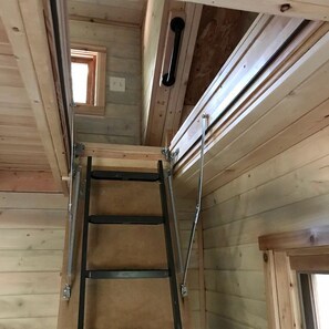 Un interior de madera con una escalera metálica, vigas de madera y un pequeño ventanal.