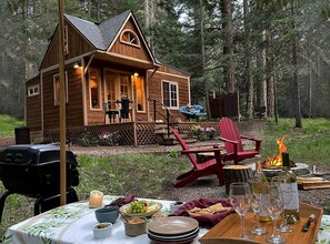 A wooden cabin with a porch, outdoor seating, a fire pit, and a table set for a meal.