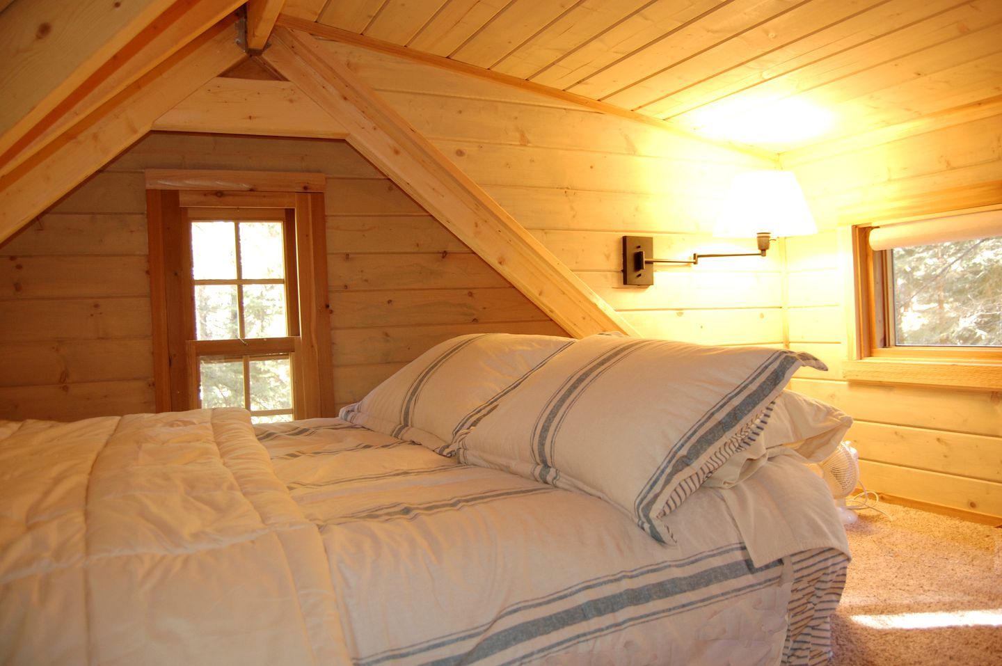 Une chambre mansardée chaleureuse, avec un plafond en pente, un lit avec une literie à rayures, une fenêtre et un luminaire fixé au mur.