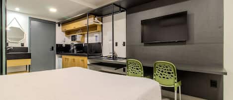 A modern hotel room with a bed, a kitchenette, a TV, and two green chairs.