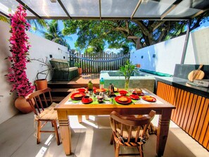 An outdoor dining area with a table set for two, surrounded by chairs, a potted plant, and a wooden fence. - Get Sense (João Pessoa)